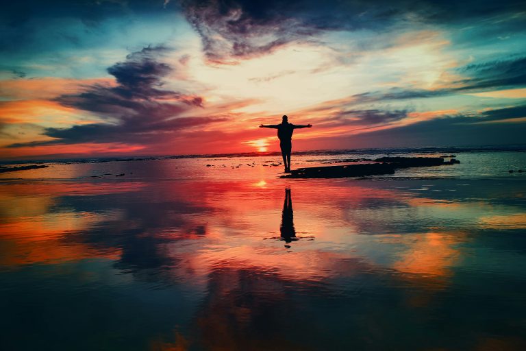 A silhouette with outstretched arms against a vibrant sunset reflected on watery sand.