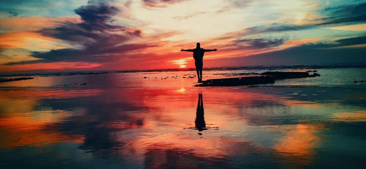 A silhouette with outstretched arms against a vibrant sunset reflected on watery sand.